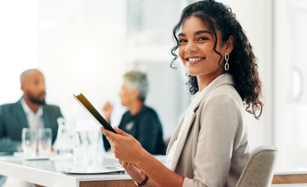 A smiling female professional with curly hair holds a digital tablet at a modern boardroom table, representing career advancement and professional training at an RTO.
