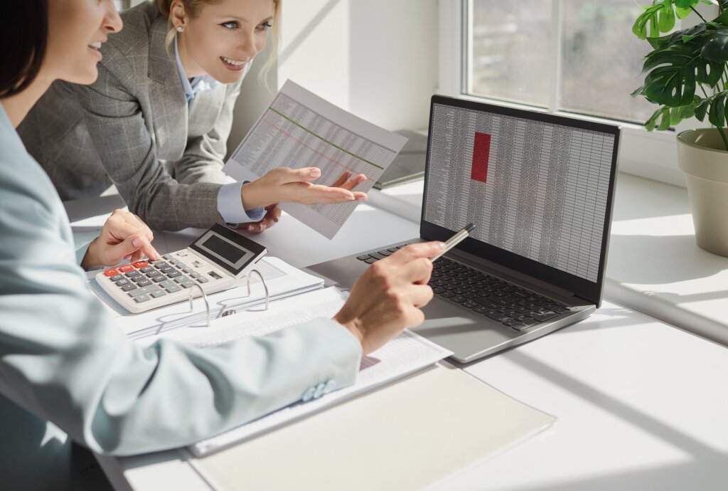 Two women in blazers work together at a white desk, with one pointing a pen at a spreadsheet on a laptop screen while the other looks on holding a printed document next to a desktop calculator.