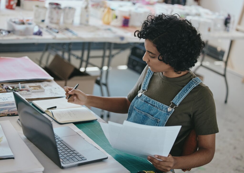 A business administration professional manages office paperwork and cross-references documents with an open notebook and laptop to keep operations running smoothly.