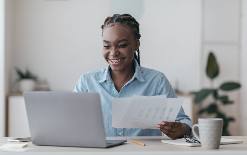 A smiling student reviews her coursework feedback on a laptop while holding printed assessment documents at her desk.