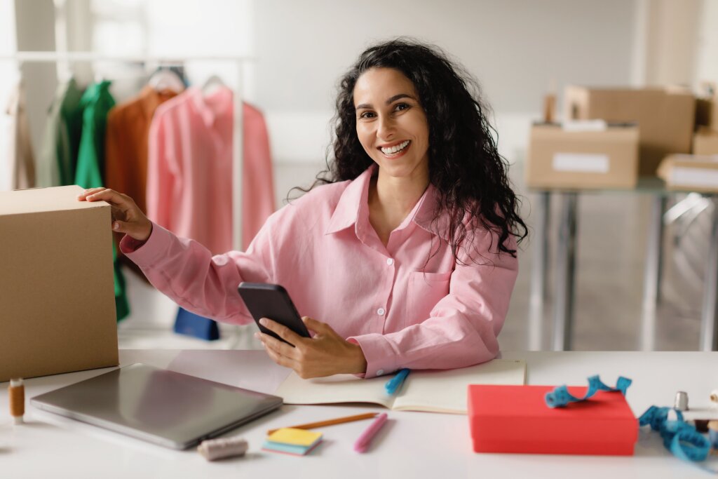 A smiling woman with long curly hair wears a pink button-down shirt and sits at a white desk holding a smartphone, with a cardboard shipping box, laptop, and office supplies in front of her, and clothing racks and packages in the background.