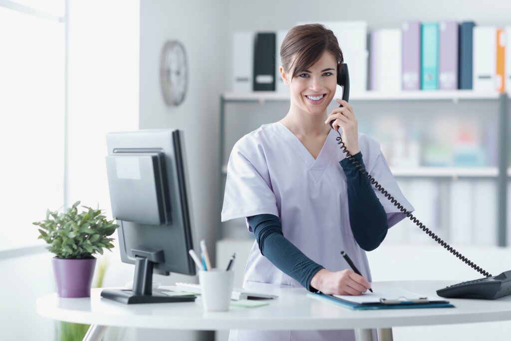 A smiling woman in light grey scrubs talks on a desk phone while writing on a clipboard next to a computer monitor.