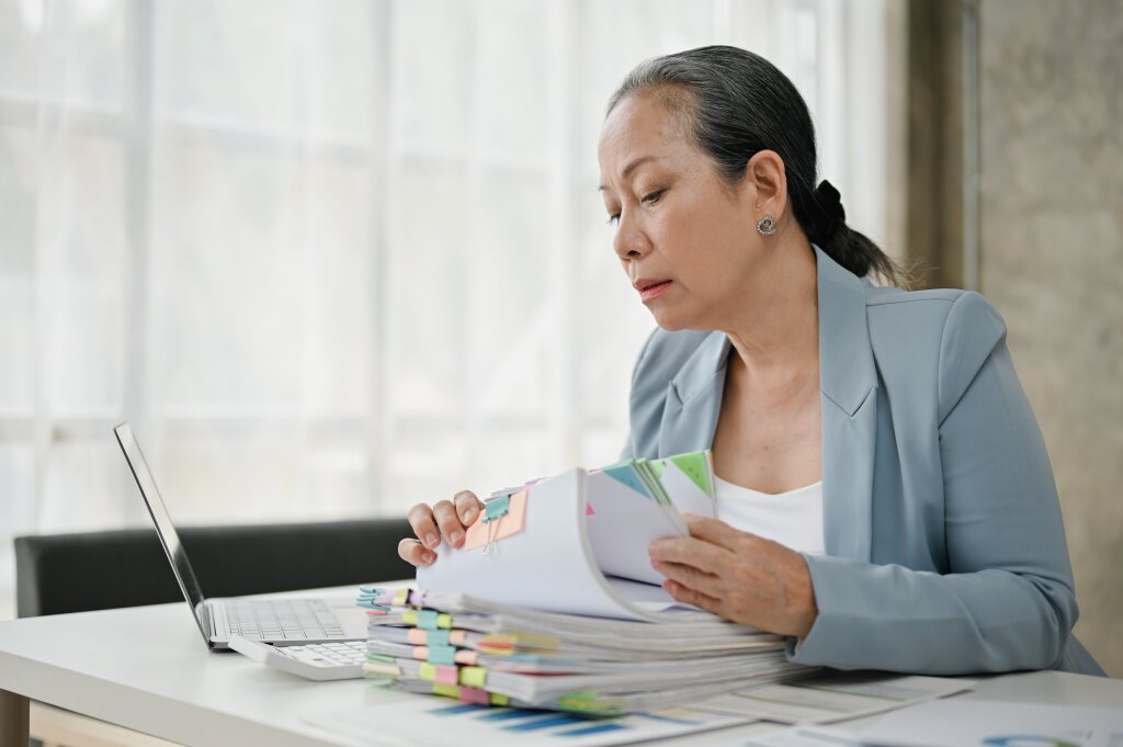 An experienced records officer meticulously reviews and categorises physical corporate archives alongside digital database files at her office workstation.