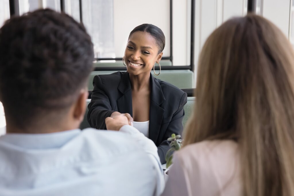 A medium shot from behind a couple as a smiling Black woman in a black blazer and hoop earrings shakes hands with the man after helping them with a financial matter.