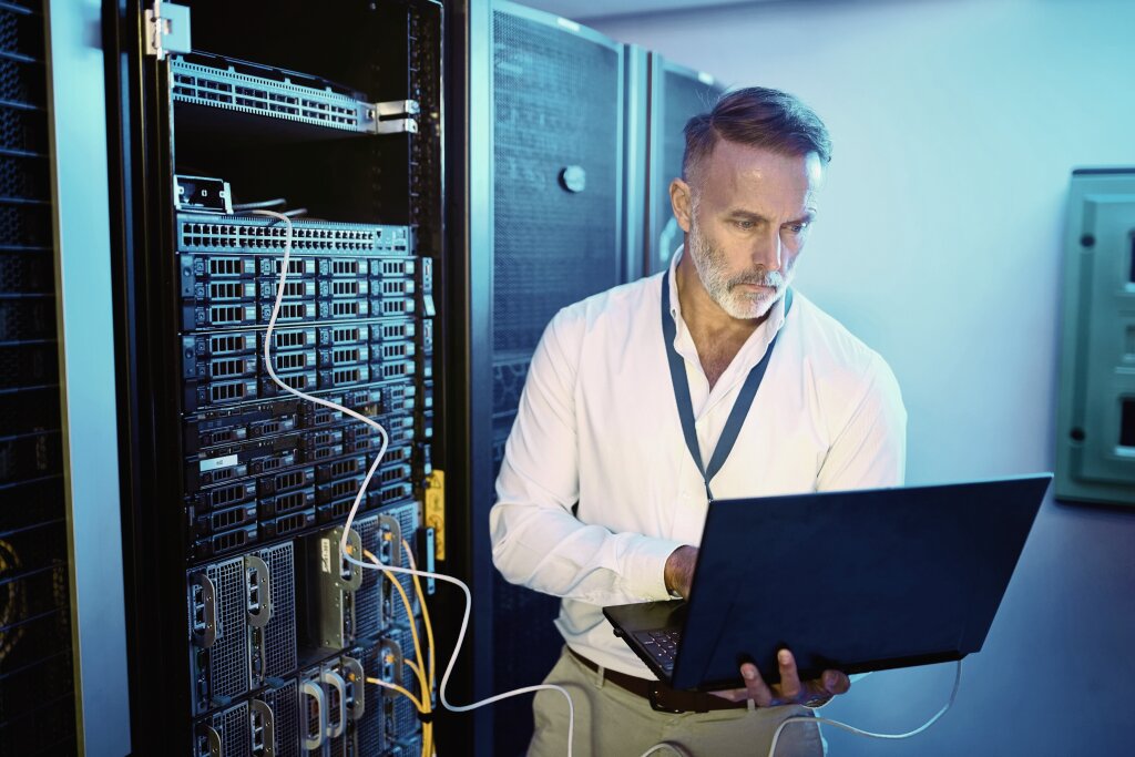 A focused IT professional or network engineer monitors server performance and configures system infrastructure using a laptop inside a data center server room.