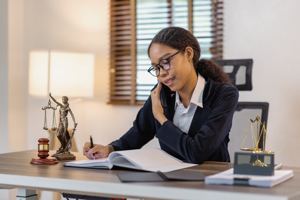 A young woman with glasses and a black blazer sits at a desk, talking on a smartphone while writing in a large open book, with a Lady Justice statue and small scales of justice nearby.