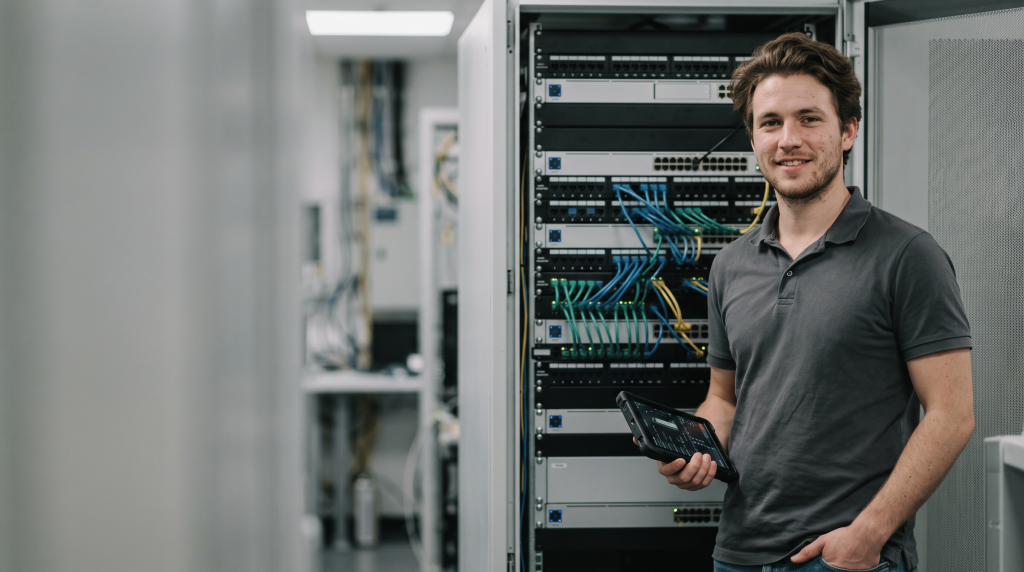A student in a grey polo shirt stands smiling next to an open network server rack, with one hand in their pocket and the other holding a digital tablet.