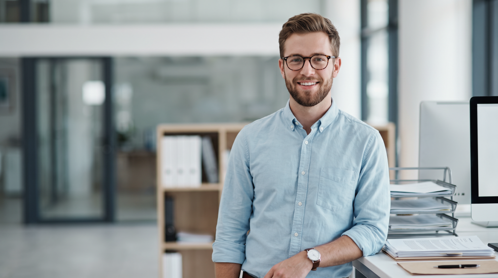 A confident project management student or junior project coordinator stands ready to organize project deliverables and manage team workflows at his office workstation.