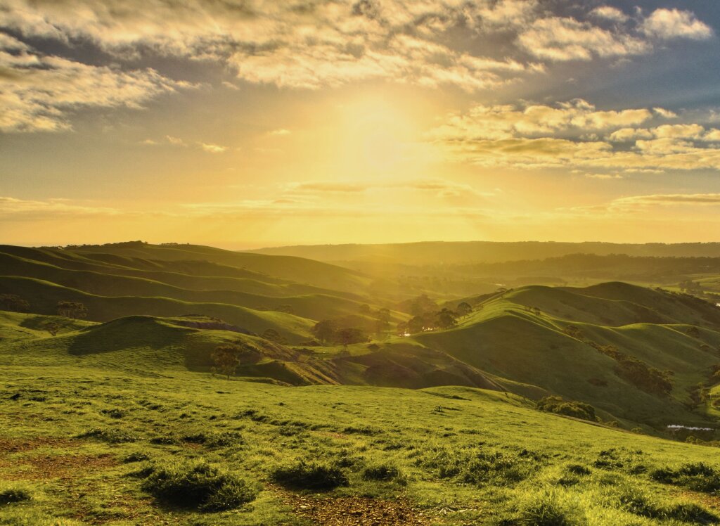 Sunlight streams down over lush, rolling green hills and valleys under a partly cloudy sky at sunrise or sunset.