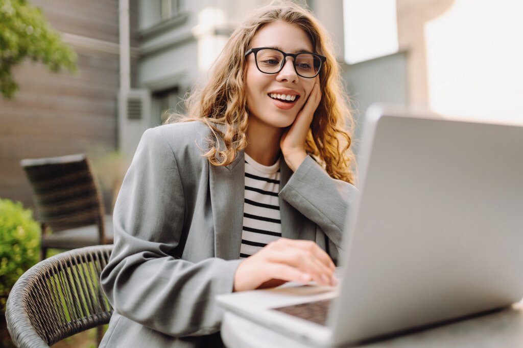 A smiling student wearing glasses reviews her online course modules on a laptop while studying outdoors at a cafe table.