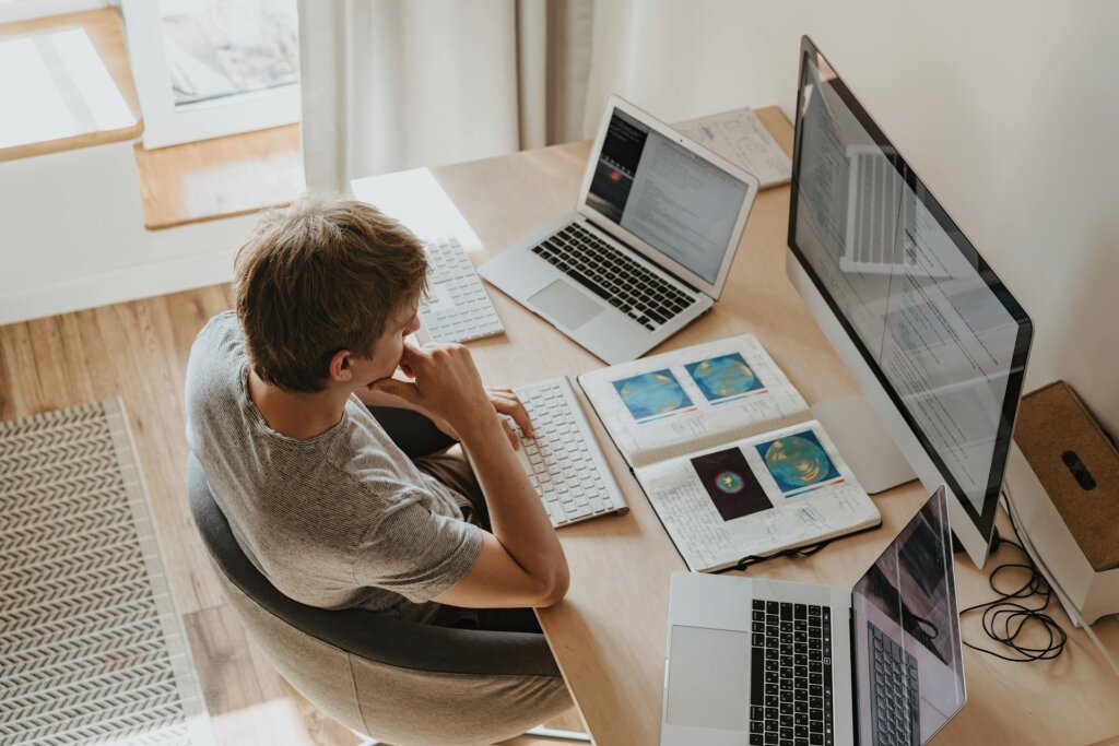 A focused student sits at a home desk with multiple computer screens and open textbooks, deeply engaged in remote study.