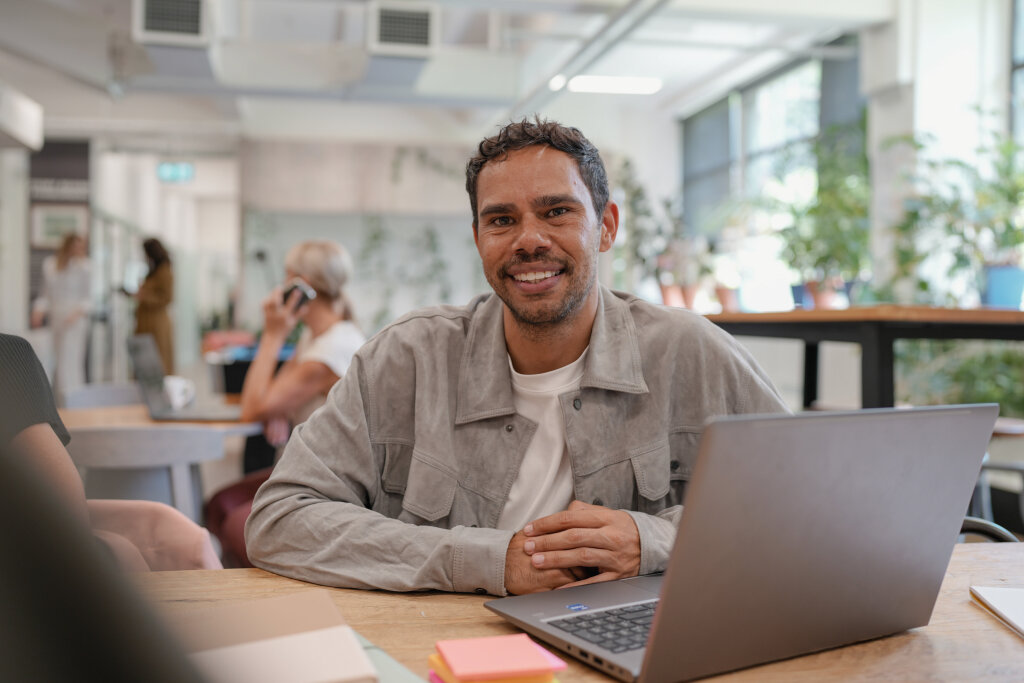 A smiling First Nations student sits at a timber desk with a laptop, engaging in online learning inside a collaborative workspace.