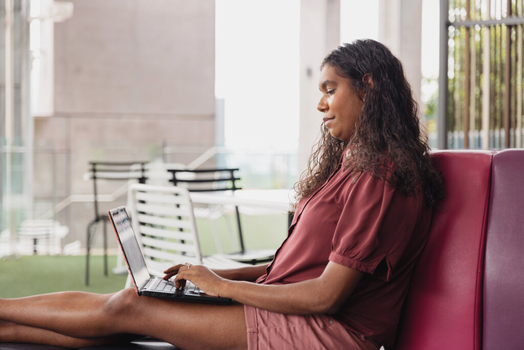 A First Nations trainee reviews a digital training module on her laptop while working in a modern, open-air office breakout area.