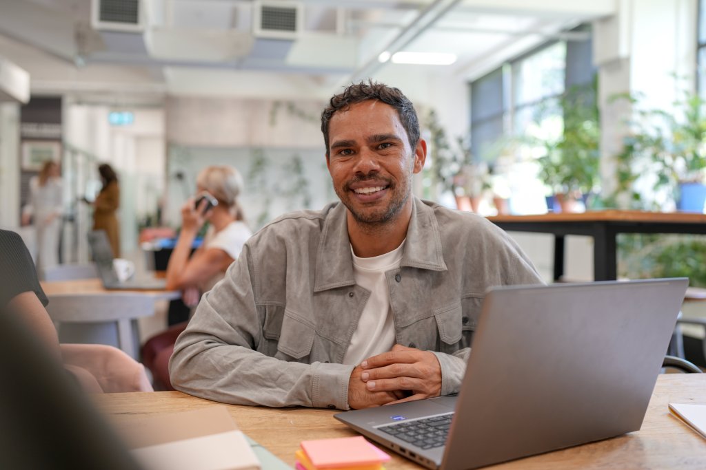 A smiling First Nations adult student sits at a collaborative workspace with a laptop, engaging in online learning or flexible study.