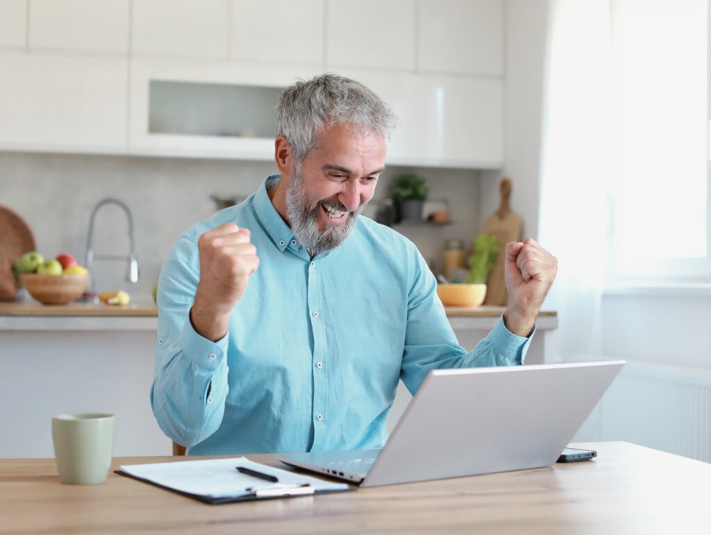 A mature-aged online student celebrates with fists raised at a desk after successfully finishing his vocational course assessment.