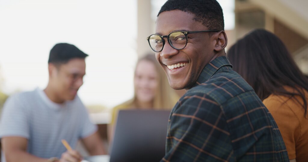 A student smiles confidently over his shoulder while studying with a laptop alongside classmates.