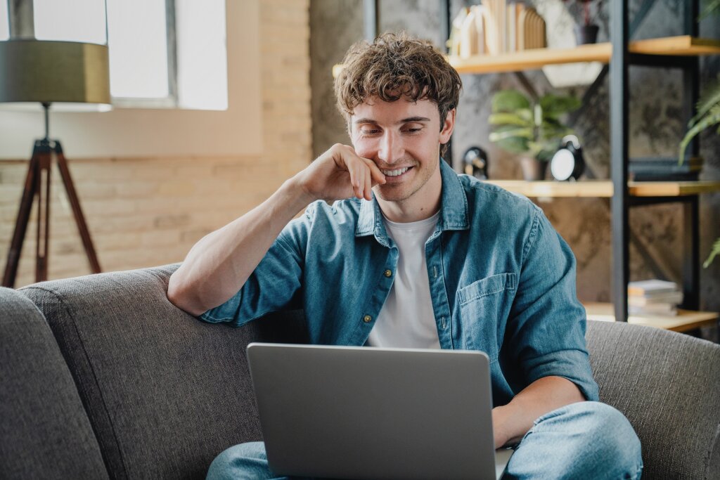 A smiling student sits comfortably on a couch while using his laptop to read through the online student handbook.