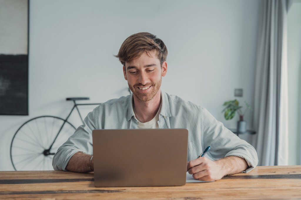 A smiling online student takes notes with a pen while studying course materials on his laptop during a flexible learning session.
