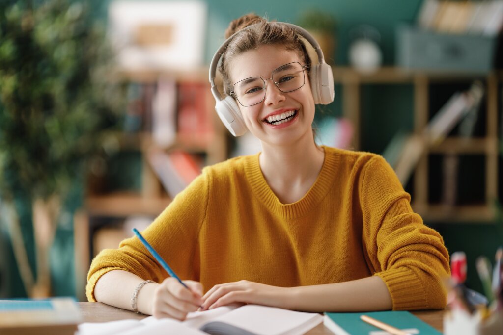 A smiling student wearing headphones writes in a book while studying her online course materials at home.