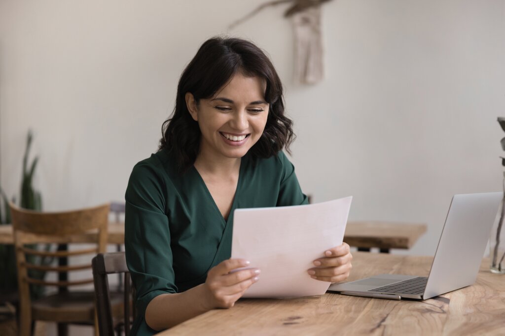 A smiling student reviews her approved government funding letter at a wooden desk with an open laptop nearby.