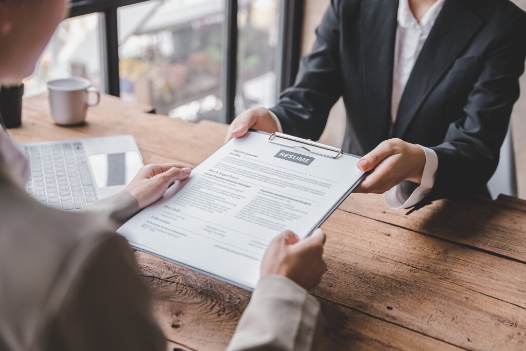 A graduate hands her newly written resume on a clipboard to an interviewer, showcasing her qualification and training.