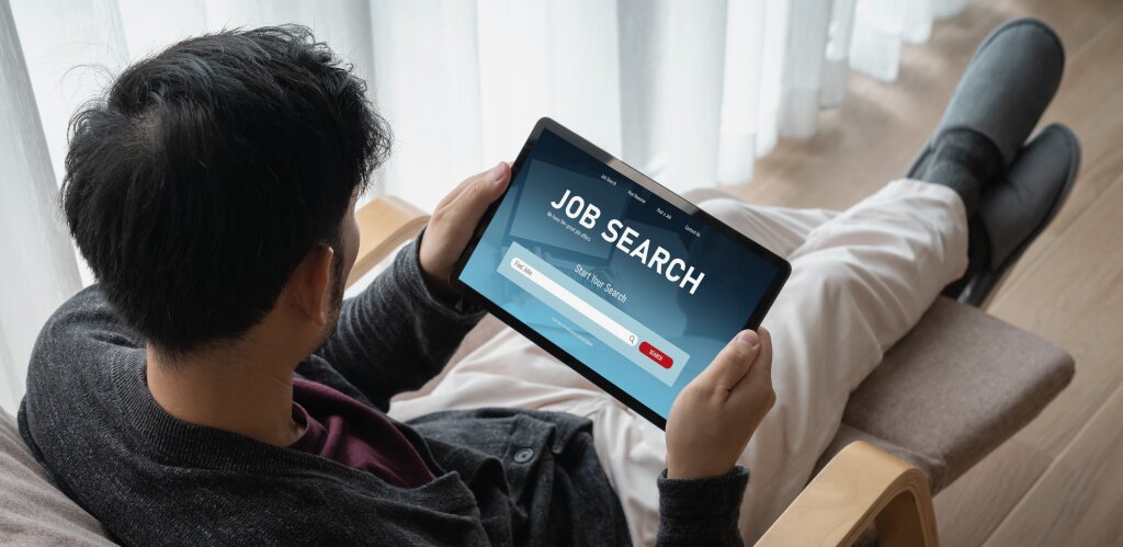 An over-the-shoulder view of a man sitting in an armchair holding a tablet displaying a "Job Search" website interface.