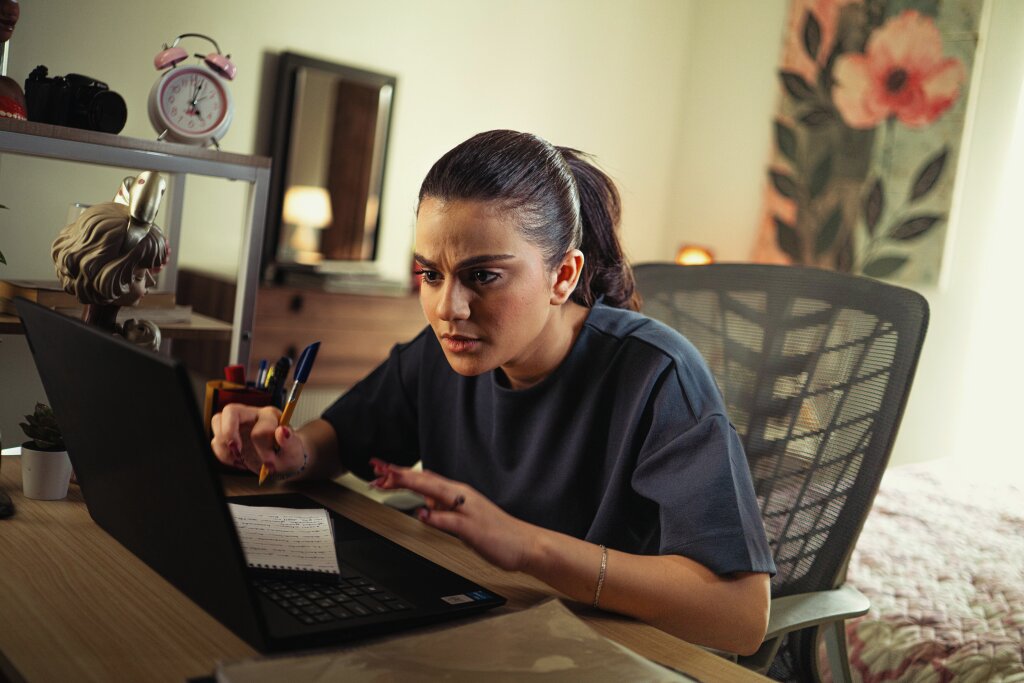 A young adult learner wearing a dark oversized t-shirt, leans toward her laptop focusing on the screen, holding a pen over a notebook in a home environment.