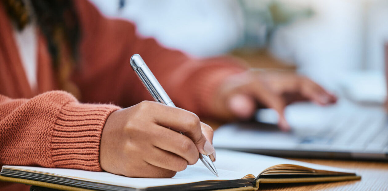 Close up of a student's hands writing in a notebook and typing on a laptop