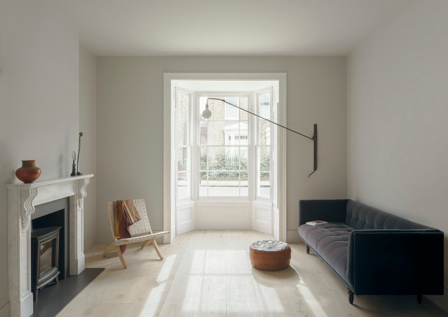 cozy, minimalist living area with modern light fixture, grey sofa, leather ottoman, and woven leather chair, white bay windows with a street view