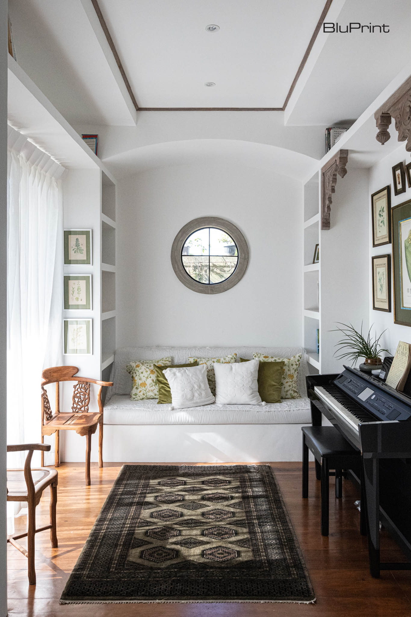 A cozy sitting area with an antique wooden chair, black upright piano, and a white sofa filled with throw pillows under a round window.