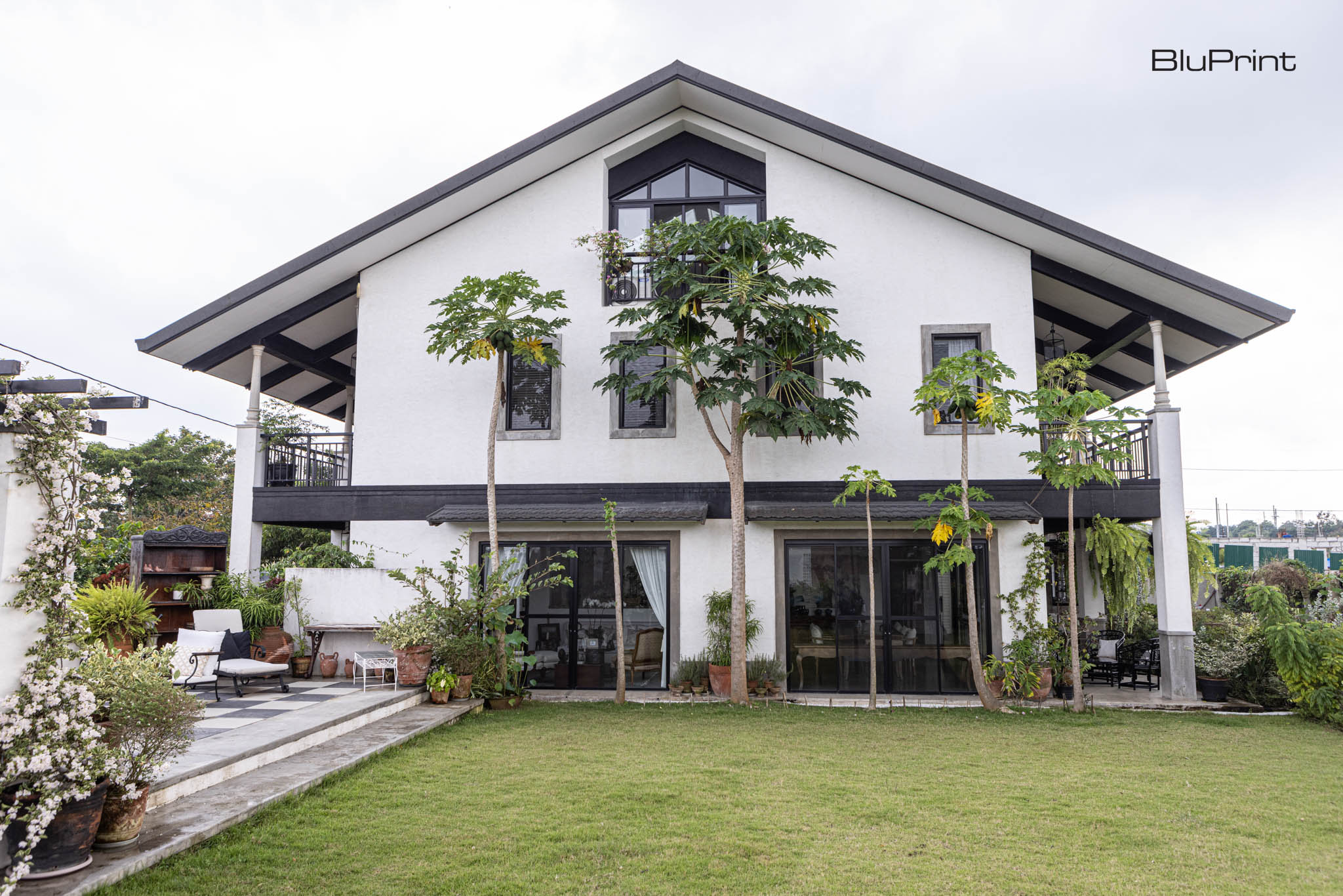 A two storey white house with pitched roof with tall trees in front, overlooking a large green lawn.