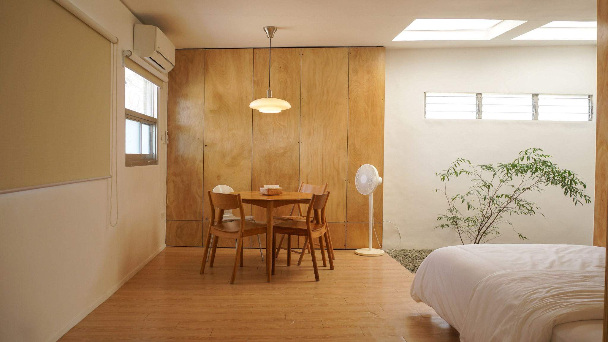 A round dining table with four seats under a modern pendant lamp. There is wood paneling on the wall and white stand fan.