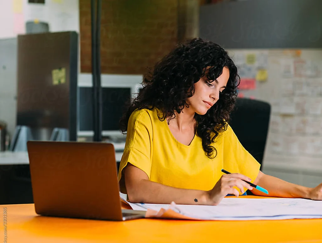 A woman with dark curly hair wearing a bright yellow top sits at a desk in an office, reviewing documents with a pen in hand next to a laptop.