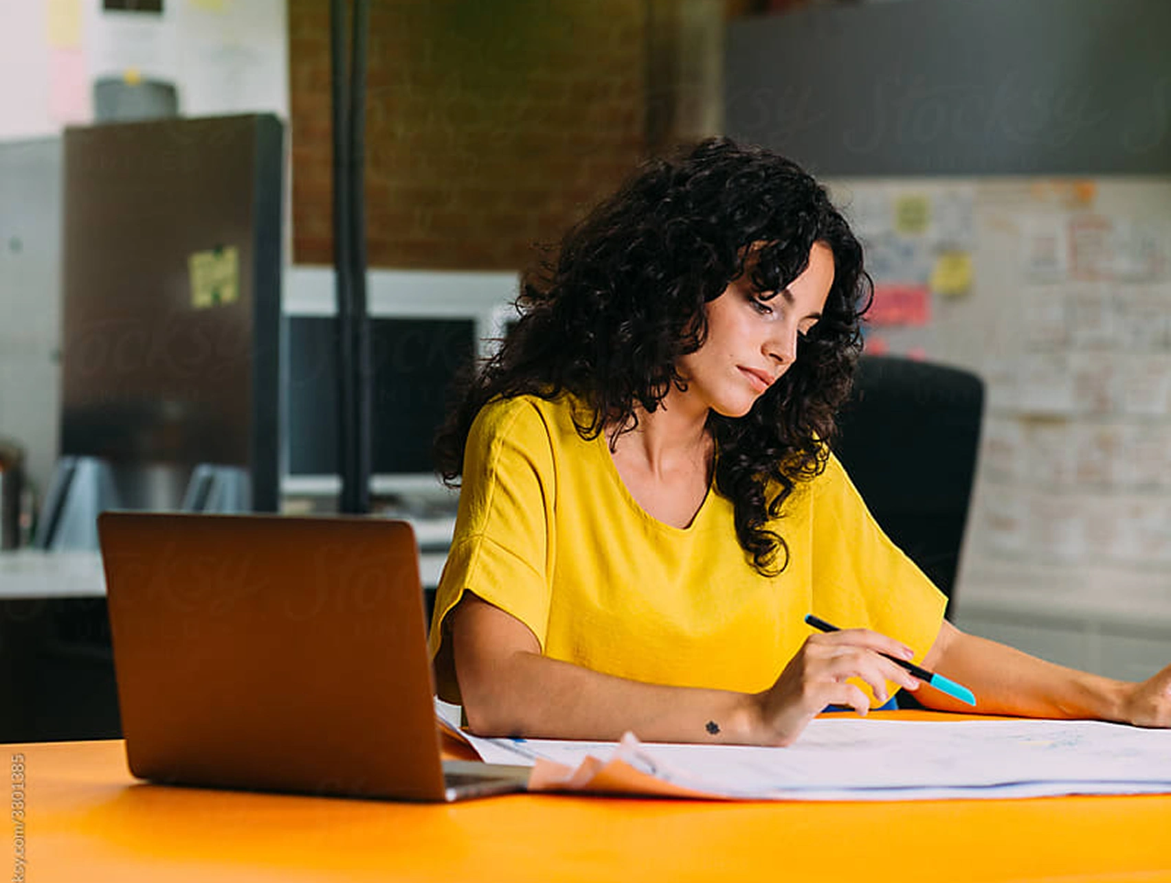 A woman with dark curly hair wearing a bright yellow top sits at a desk in an office, reviewing documents with a pen in hand next to a laptop.