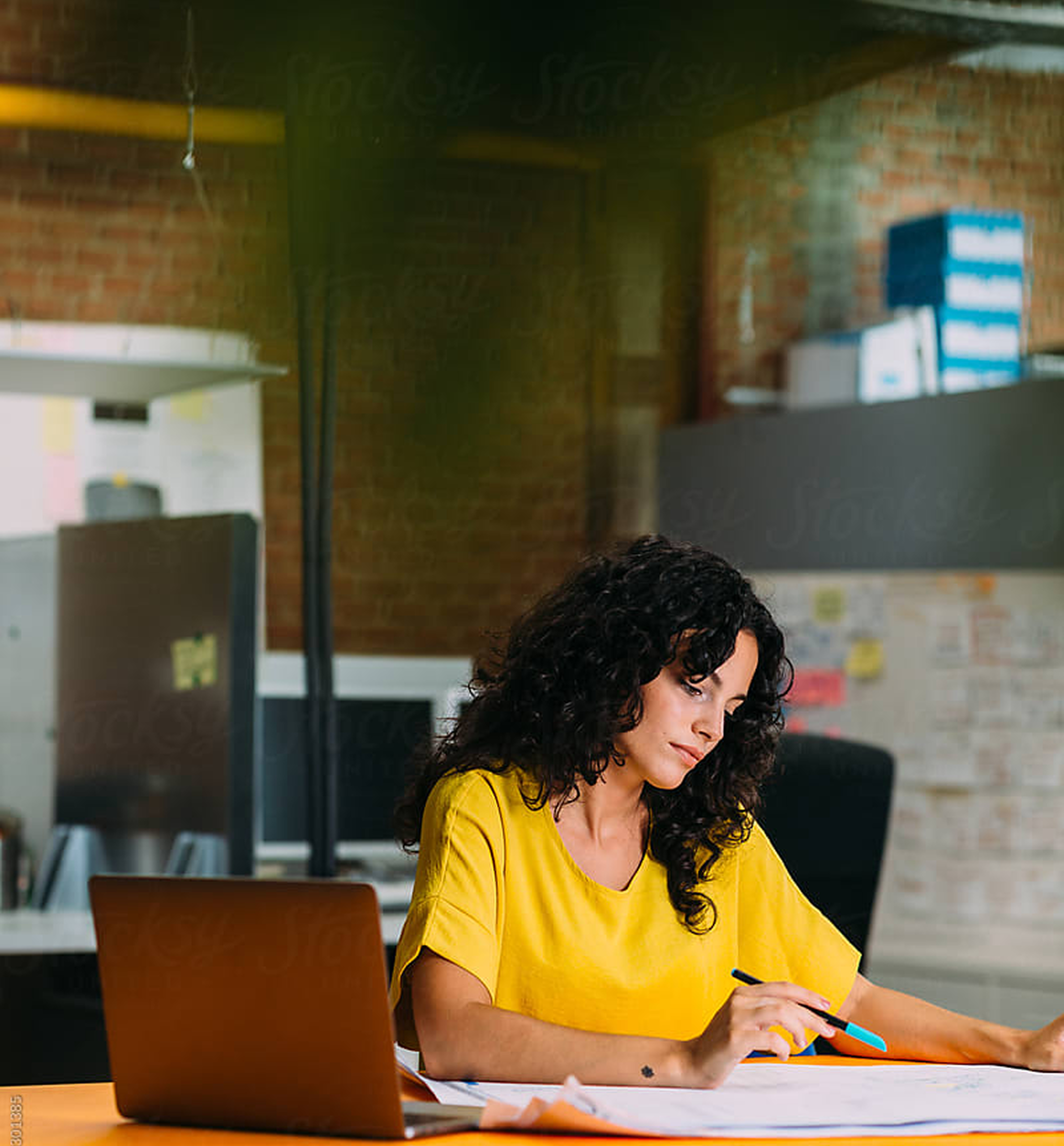 A woman with dark curly hair wearing a bright yellow top sits at a desk in an office, reviewing documents with a pen in hand next to a laptop.