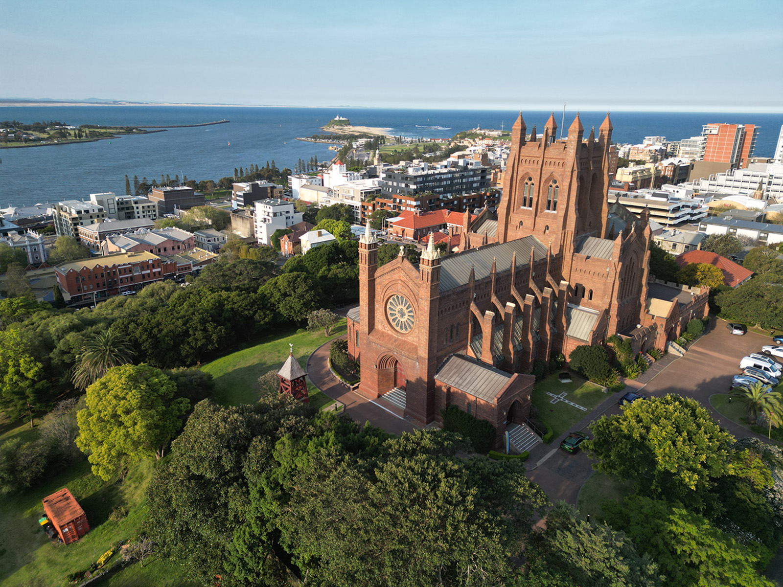 Aerial view of Christ Church Cathedral in Newcastle, Australia, surrounded by lush green trees and urban buildings, with the coastline and ocean visible in the background under a clear blue sky.