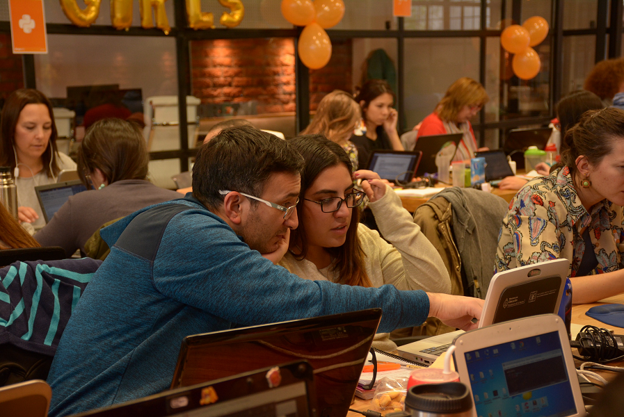 Photo of two individuals looking at laptop screen, other people in background working on laptops