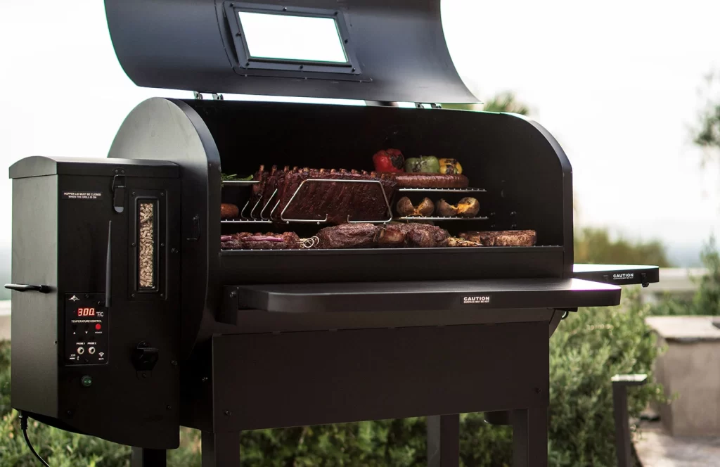 The Peak Prime by Green Mountain Grills with an open lid, showcasing grilled meats, ribs, and vegetables. The grill features a digital temperature control panel and a wood pellet hopper, designed for outdoor cooking enthusiasts.