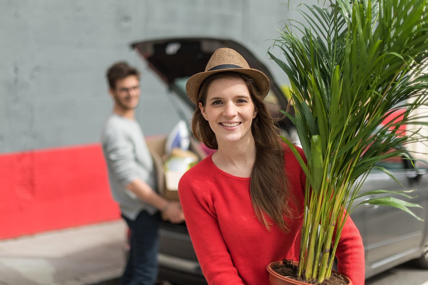 Smiling woman holding a potted plant while moving, with a man unloading boxes from a car