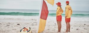 Two surf lifesavers standing next to a red & yellow patrol flag watching the beach