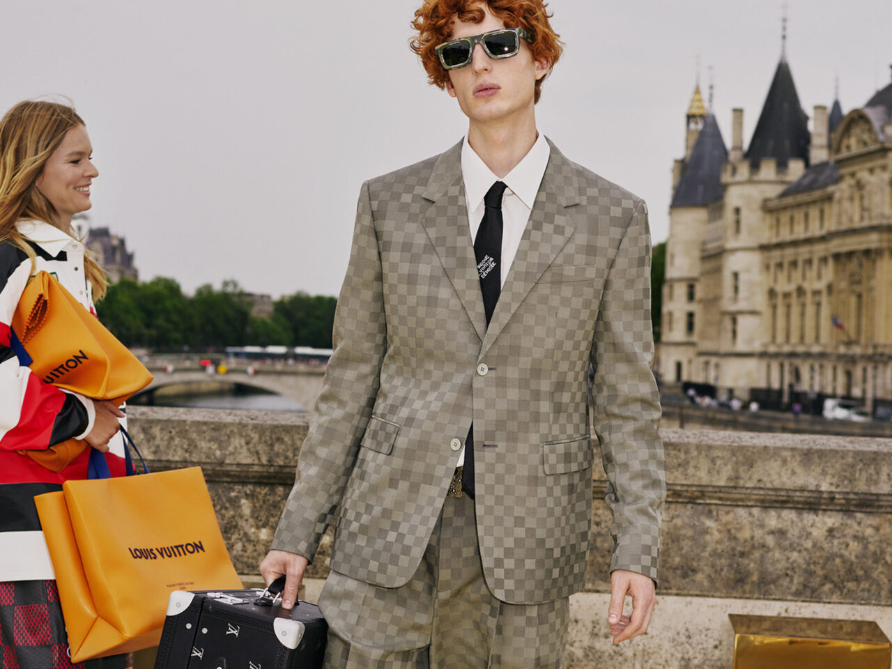 A Louis Vuitton modeling with a grey checkered suit with a trunk black Louis Vuitton bag