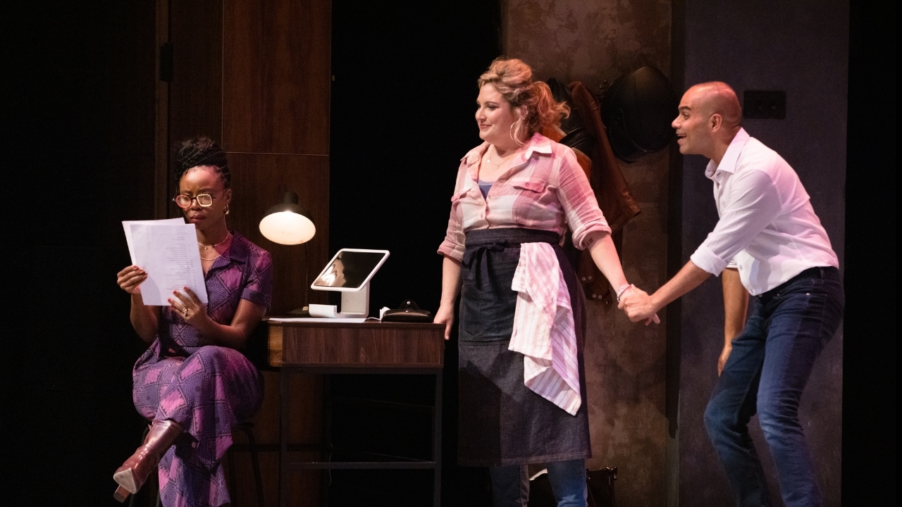 Two characters holding hands listening to a friend read a contract, on a stage in an office setting.