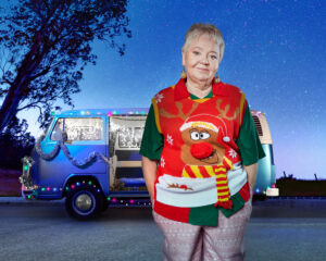 A woman with short hair wearing a Christmas jumper, standing in front of a combi van decorated in Christmas lights.