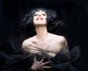 Close up of a woman with curly black hair wearing a black dress and red lipstick.