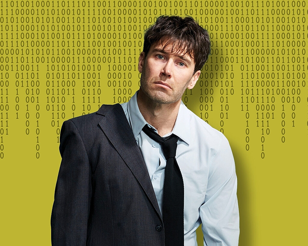 A dishevelled man with short black hair wearing a suit and tie standing against a wall of numbers.
