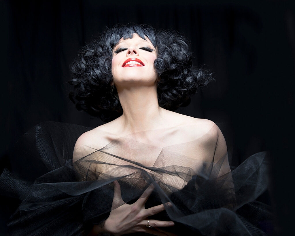 Close up of a woman with curly black hair wearing a black dress and red lipstick.