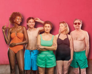 A group of five individuals wearing swimsuits smiling at each other against a pink background.