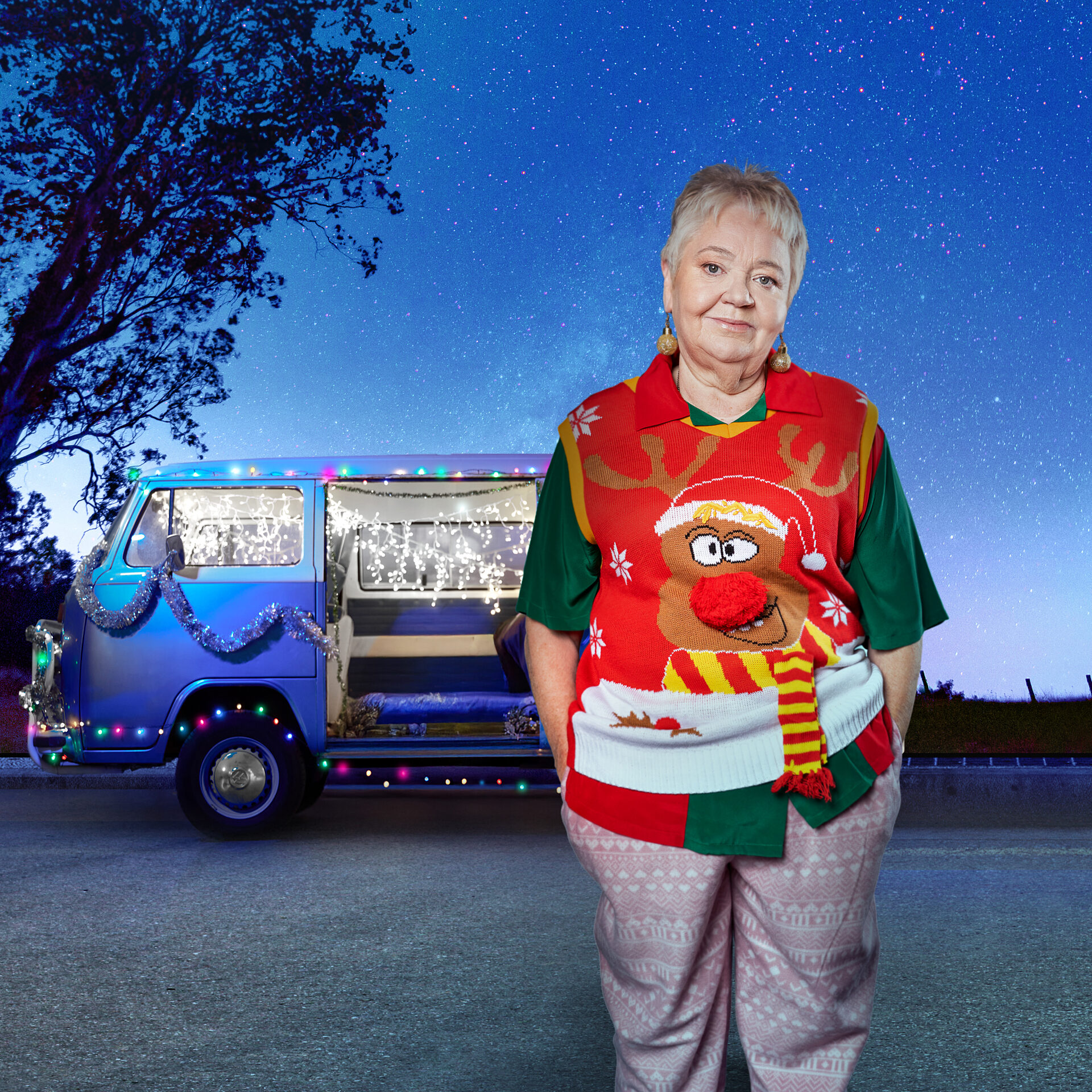 A woman with short hair wearing a Christmas jumper, standing in front of a combi van decorated in Christmas lights.