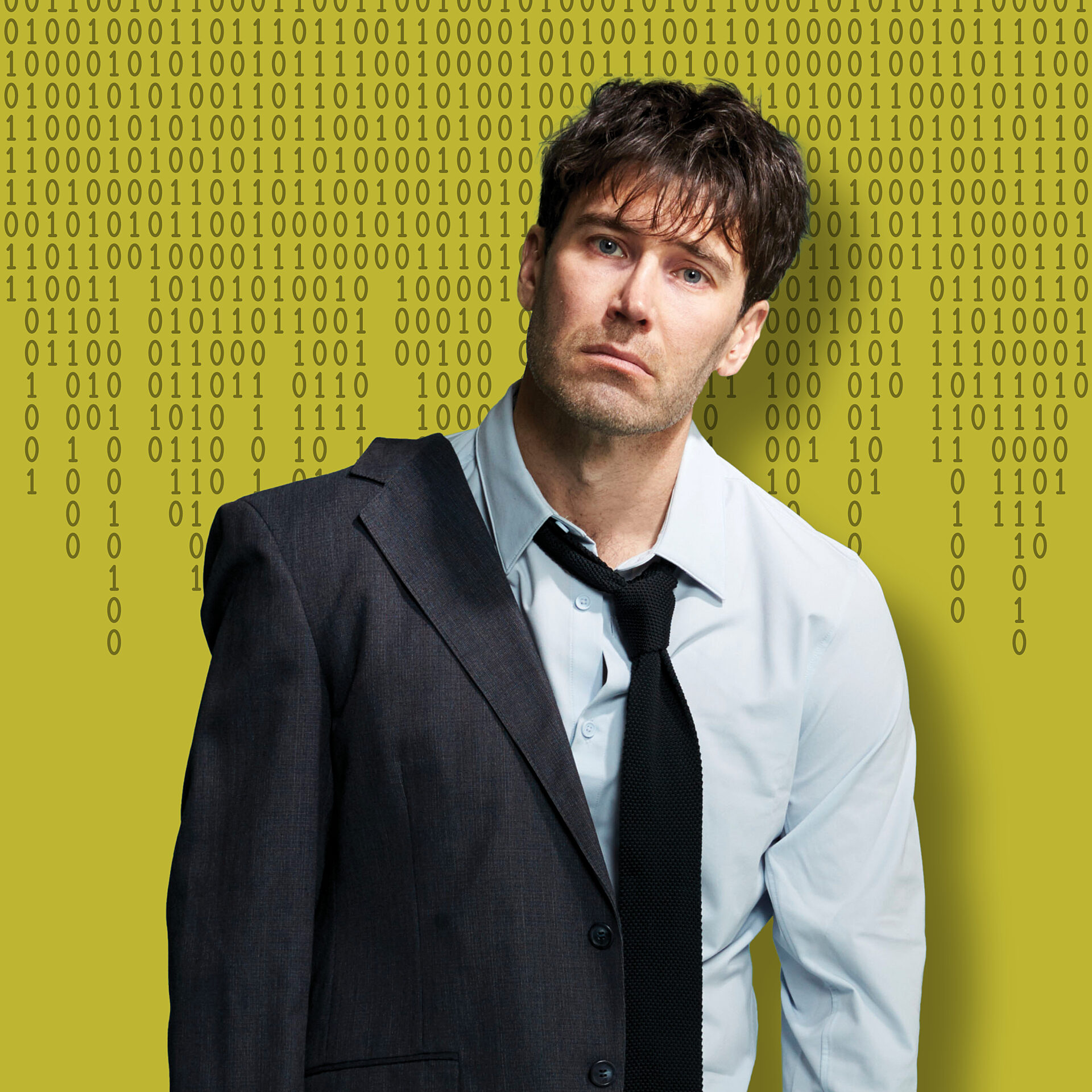 A dishevelled man with short black hair wearing a suit and tie standing against a wall of numbers.