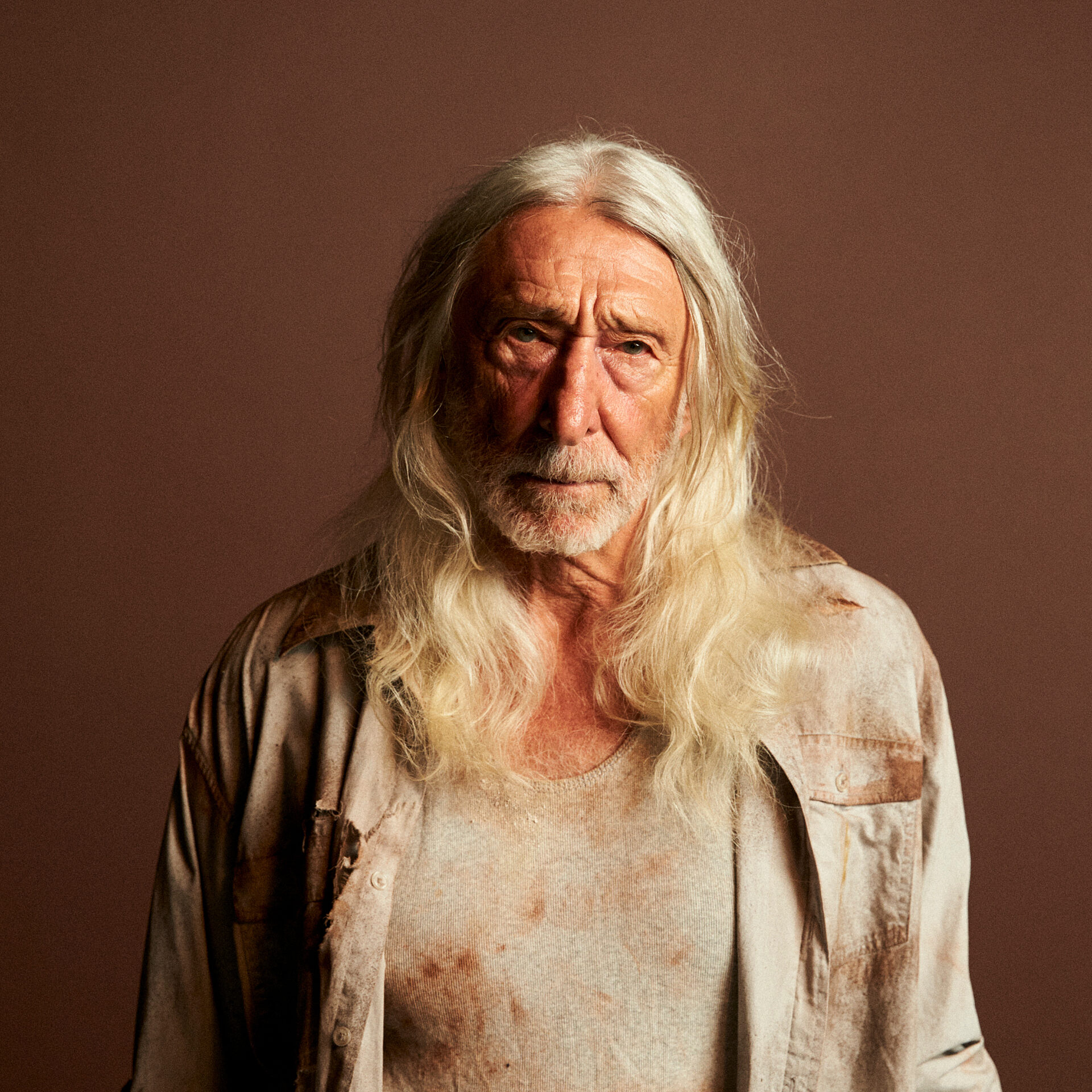 Close up of a stern old man with long white hair, wearing stained clothing.